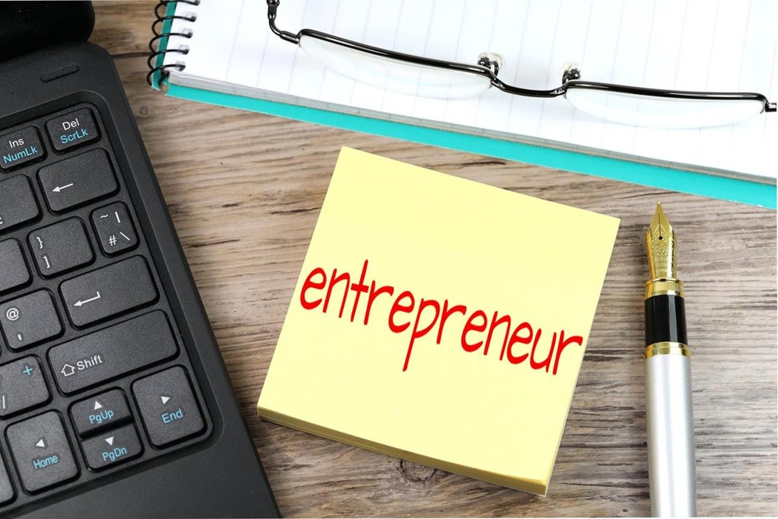 Lähikuva keltaisesta muistilapusta, johon on kirjoitettu punaisella musteella sana 'entrepreneur' toimistotarvikkeiden keskellä.