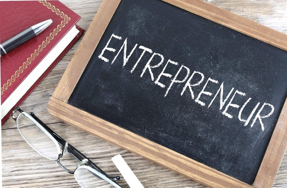 Pieni liitutaulu, jossa lukee sana 'ENTREPRENEUR', ympäröitynä toimistotarvikkeilla puisella pöydällä.